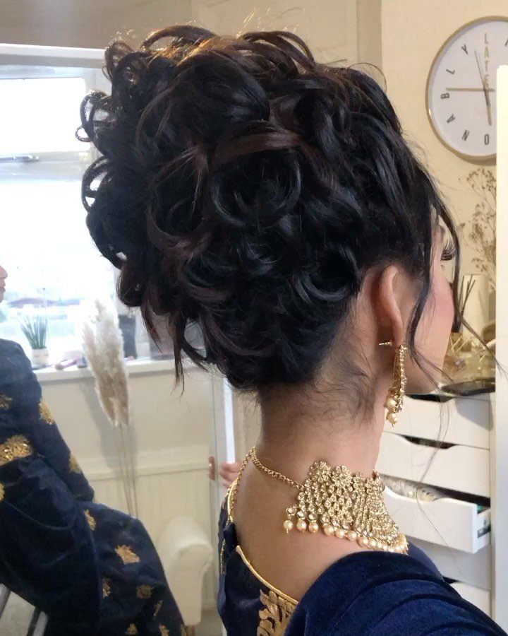 Woman with a voluminous curly updo, gold jewelry, and navy blue embroidered clothing.
