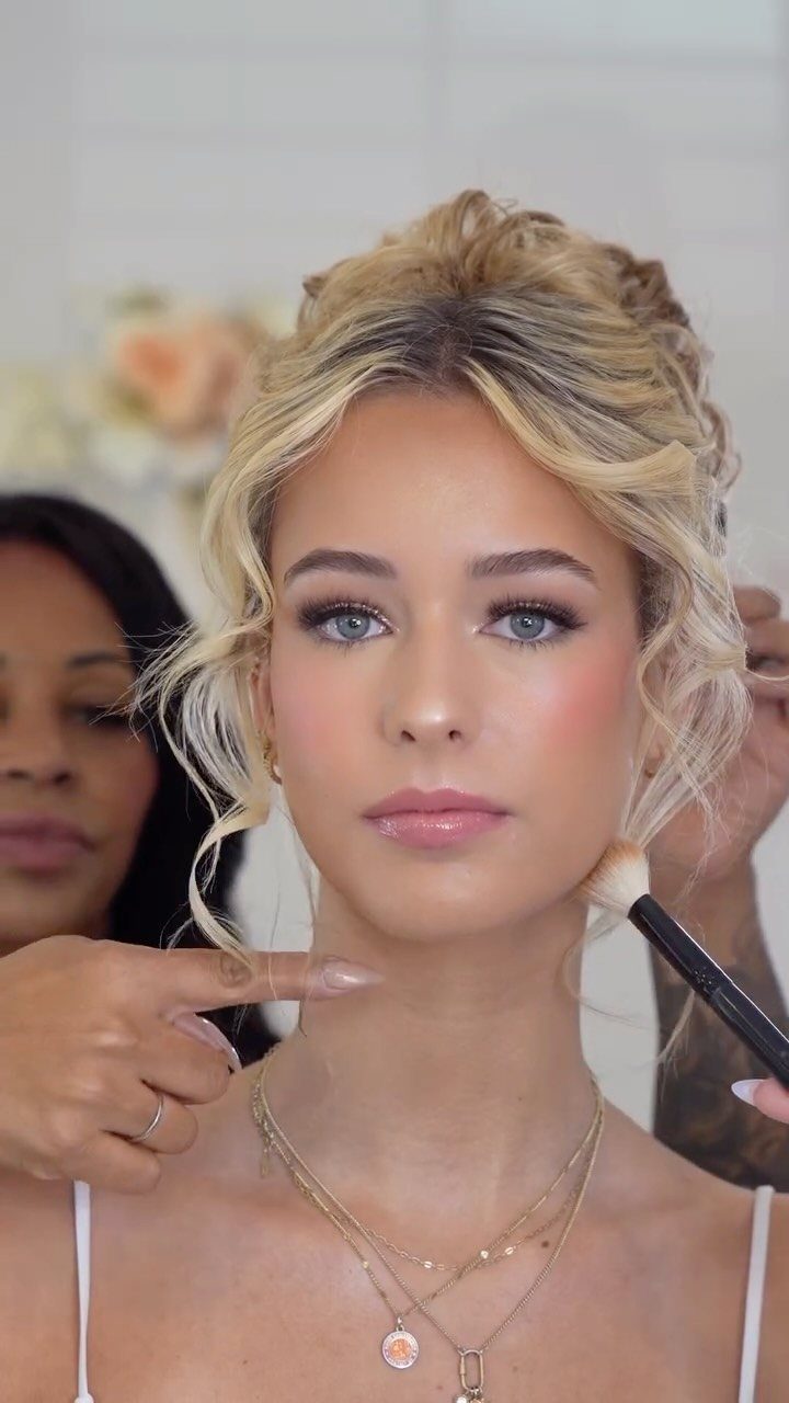 Blonde woman receiving bridal makeup application from a professional artist using a brush.