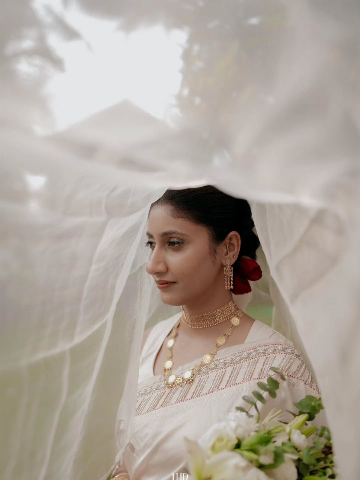 Woman with natural bridal makeup wearing a white saree and gold jewelry under a veil