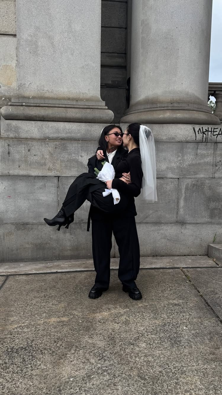 Two people wearing black suits and a bridal veil embracing by stone columns.