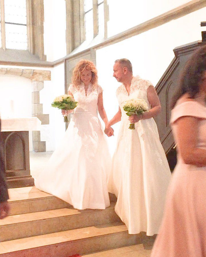 Two people in white bridal gowns holding hands and bouquets in a church