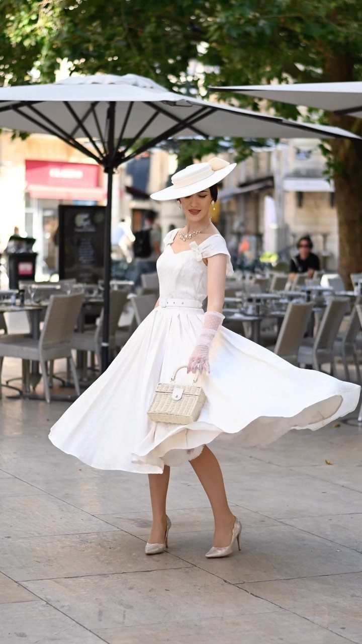 Woman in a white 1950s tea-length wedding dress and hat posing outdoors.