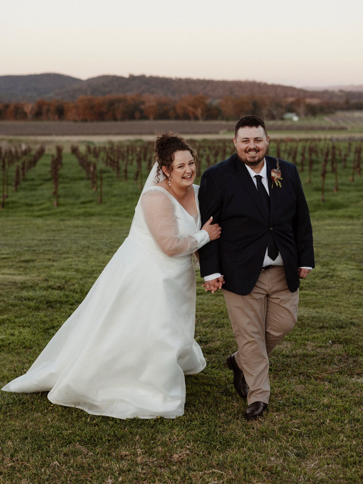 Bride and groom walking through a vineyard, groom wearing navy blazer and khaki pants.