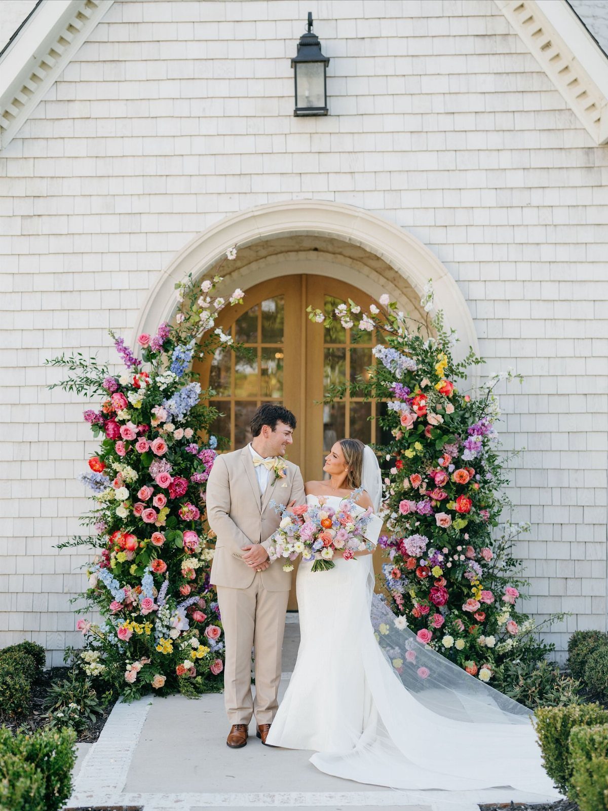 Bride and groom posing before a vibrant, multicolored floral arch at a wedding.