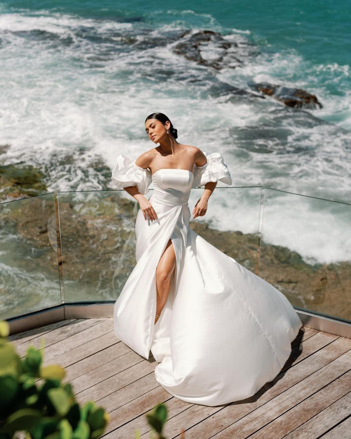 Woman in a white off-the-shoulder wedding dress posing on a deck by the ocean