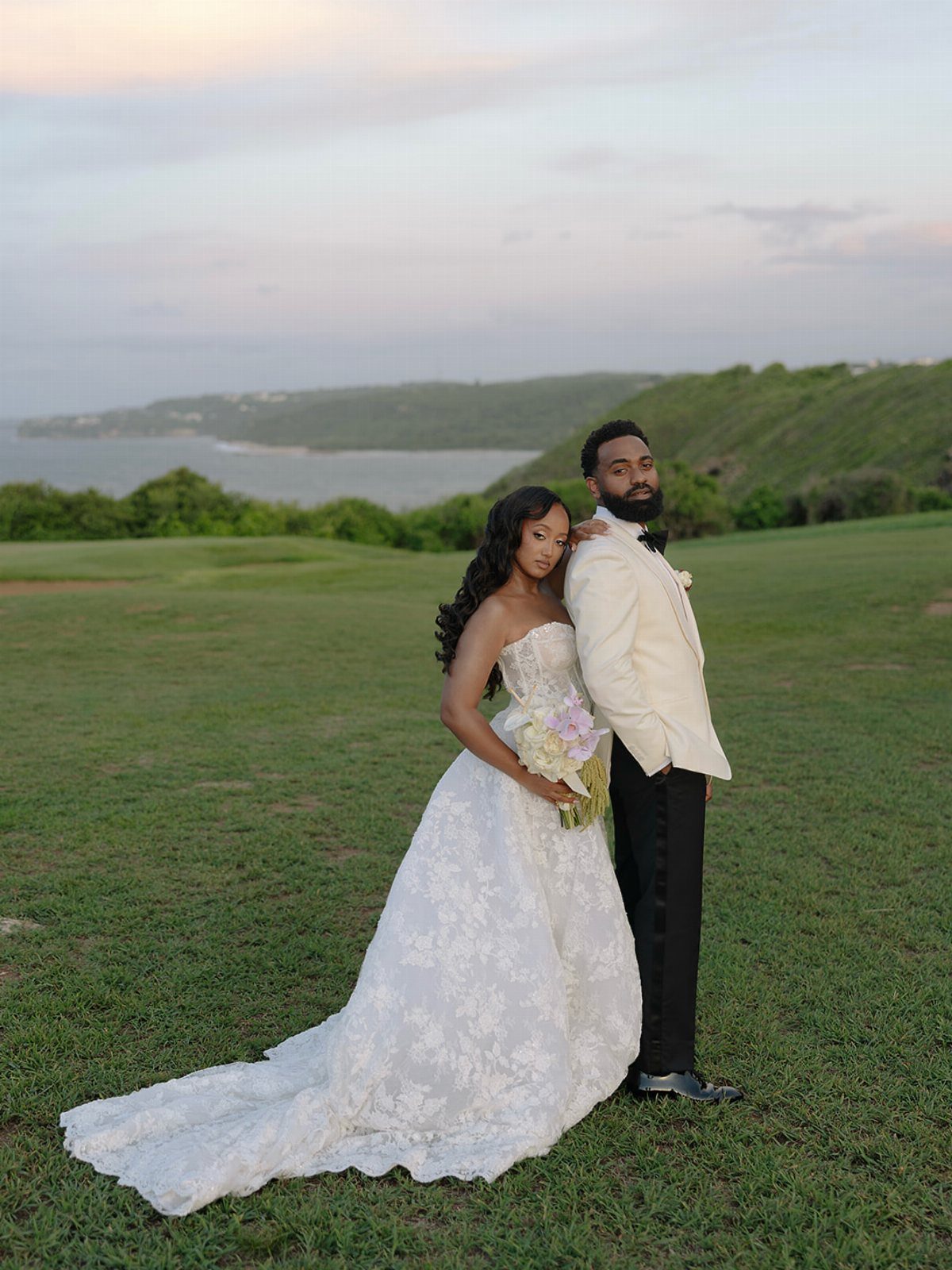 Bride in white lace wedding dress and groom in white tuxedo posing outdoors.