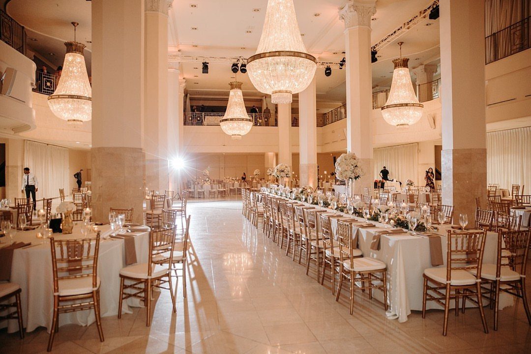 Luxury ballroom with crystal chandeliers, long white tables, and gold chairs for wedding reception.