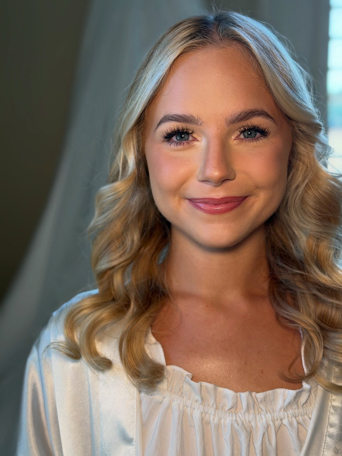 Woman with blonde wavy hair wearing a white satin robe and natural bridal makeup