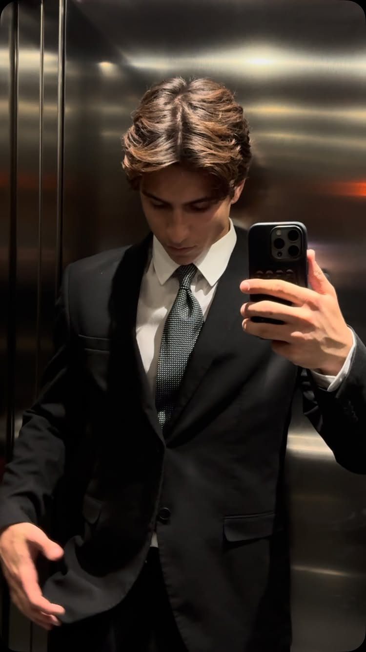 Young man in a black suit and grey tie taking a mirror selfie.