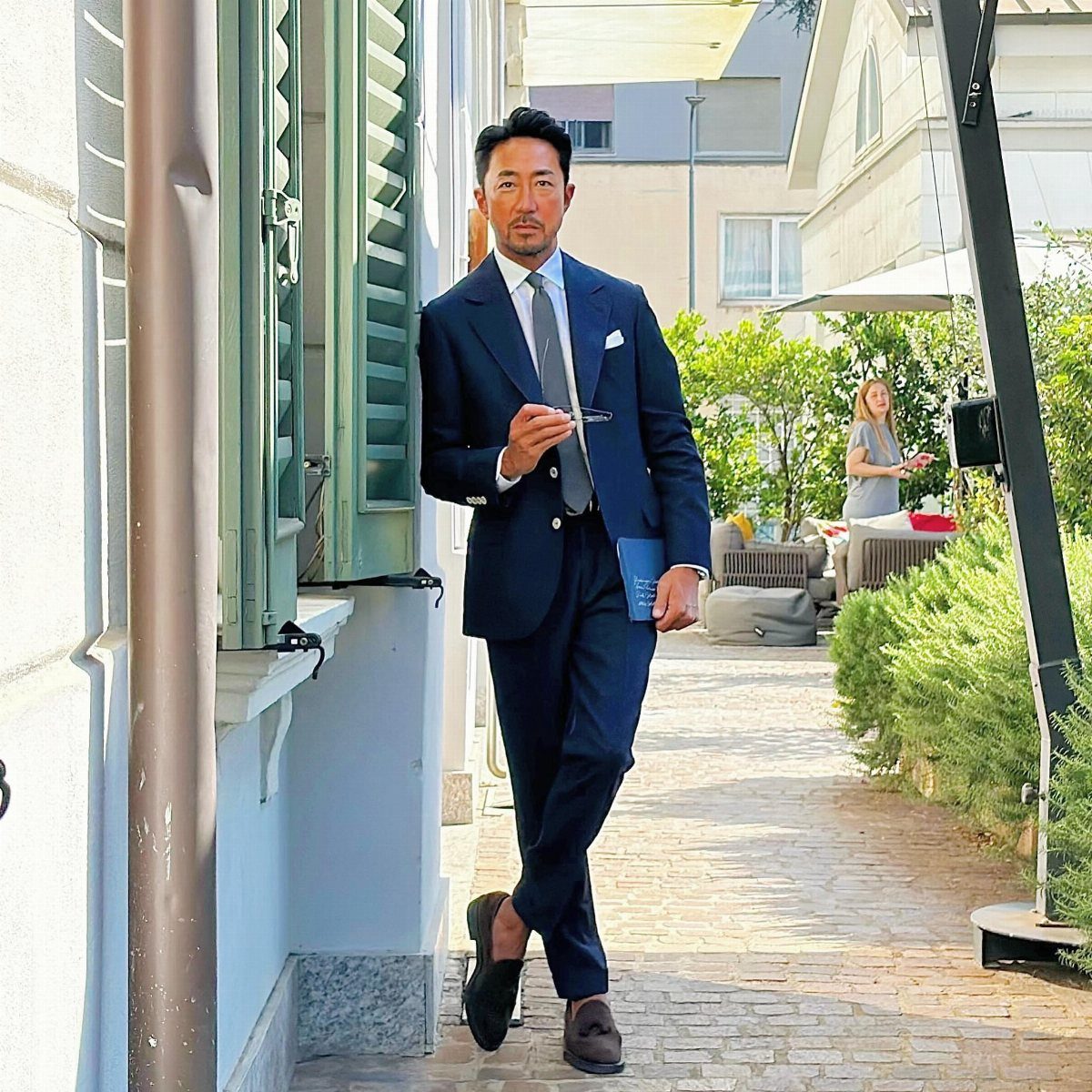 Man in a navy blue suit and grey tie leaning against white wall