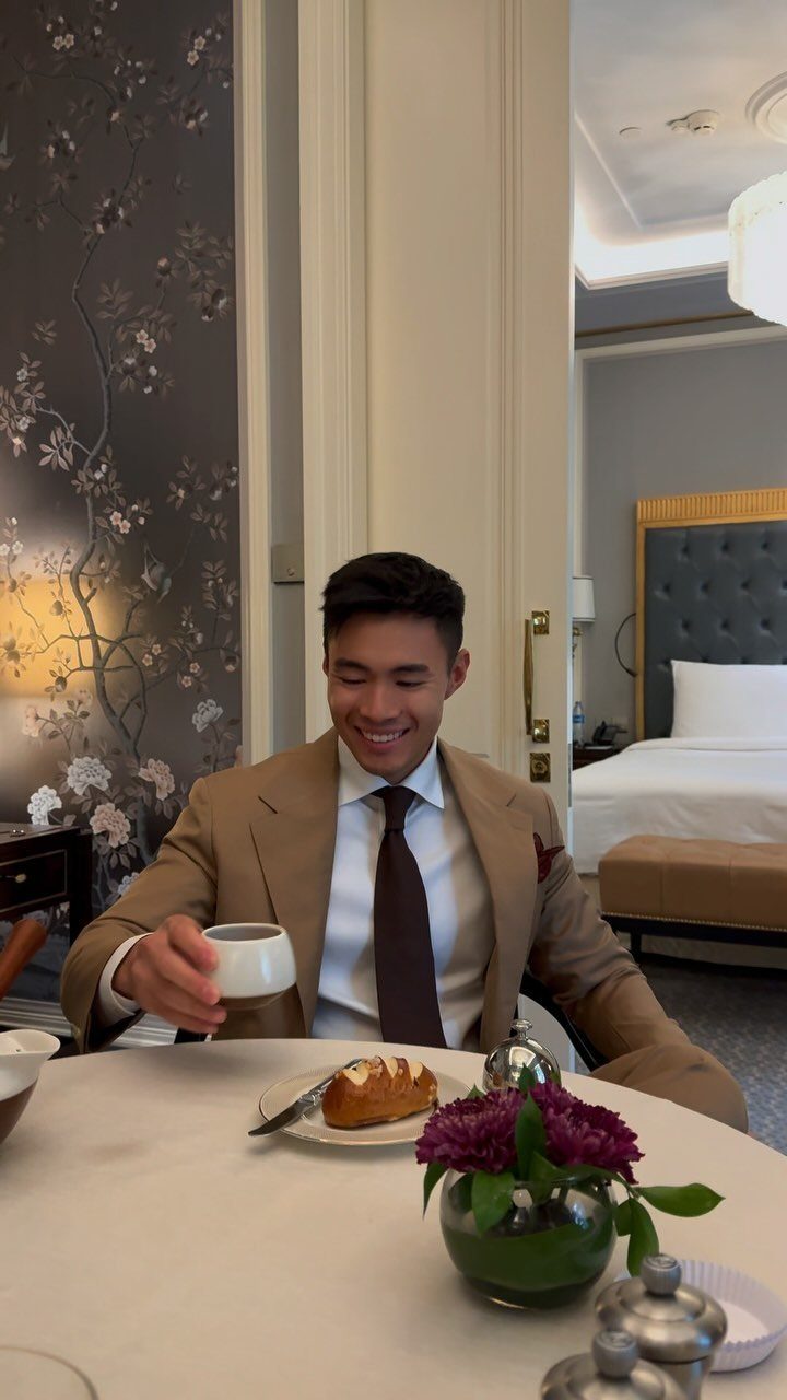 Man in tan suit and brown tie drinking coffee at a breakfast table.
