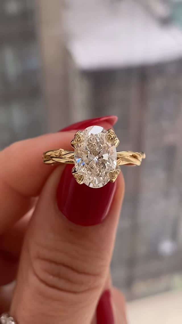 Gold engagement ring with oval diamond and vine-inspired band held by red nails