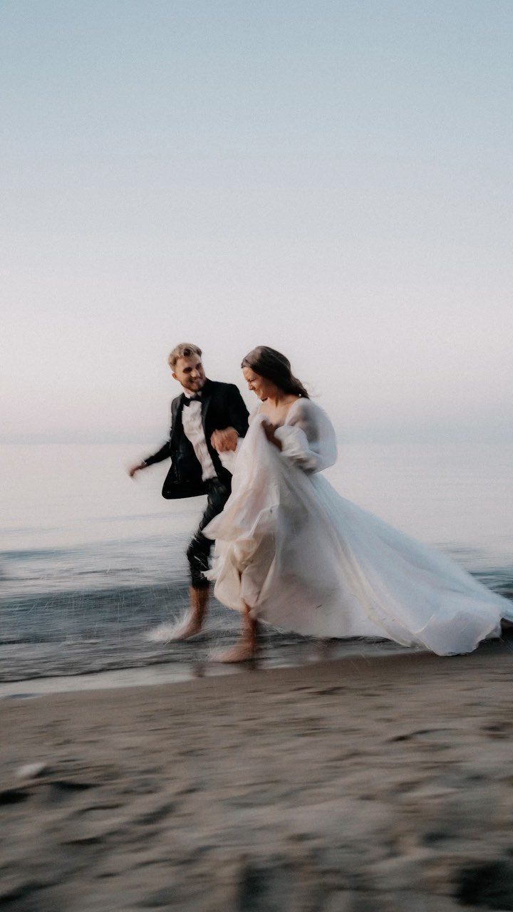 Bride in a white wedding dress and groom in tuxedo running on beach