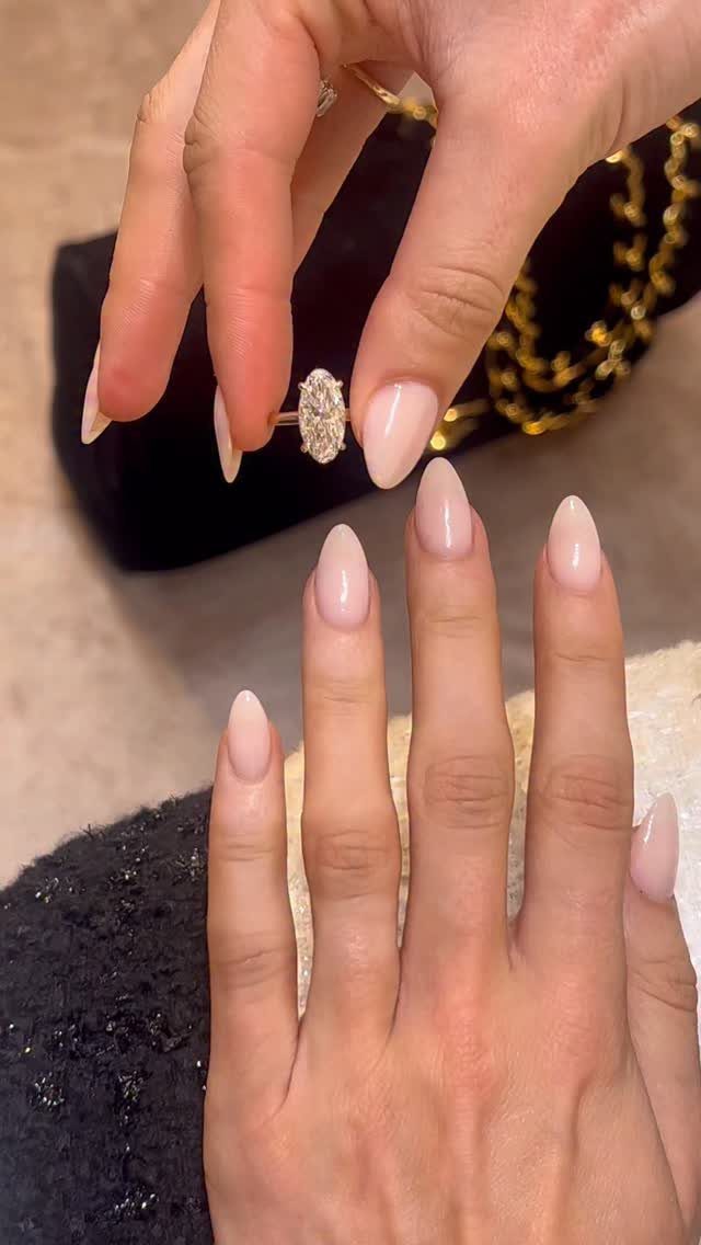 Oval diamond gold ring held between fingers above a hand with nude nails