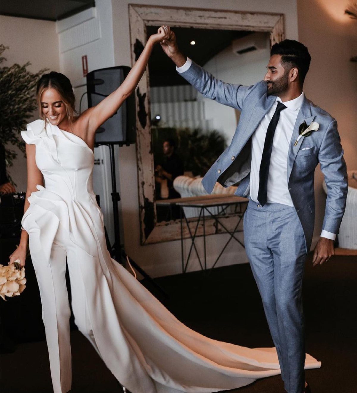 Bride in a white one-shoulder bridal jumpsuit dancing with groom in blue suit