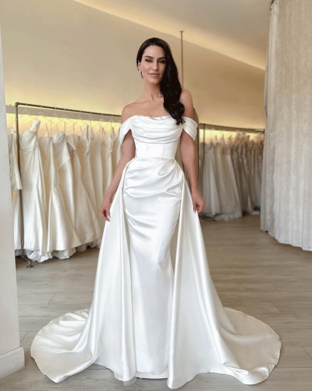 Woman in a simple white off-the-shoulder satin wedding gown standing in a bridal boutique