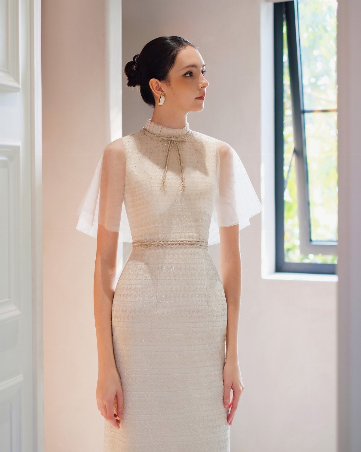 Woman in a white form-fitting wedding dress with sheer cape sleeves and gold belt