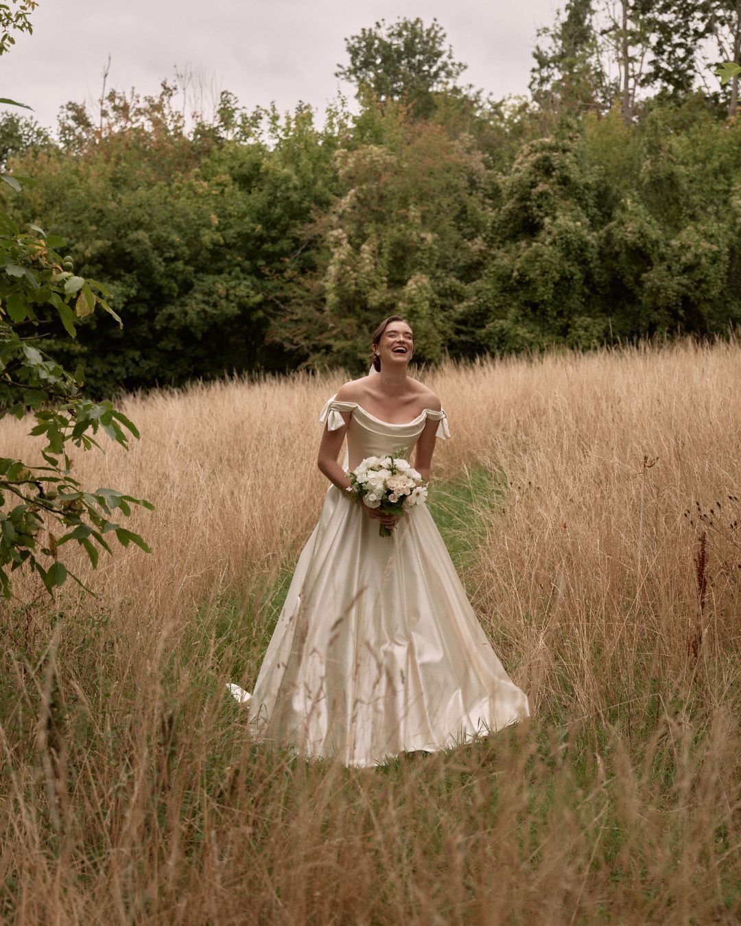 Woman in white off-the-shoulder wedding gown holding bouquet in golden grass field