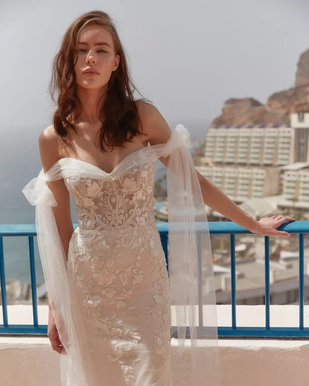 Woman in off-white off-shoulder lace wedding dress posing on a coastal balcony