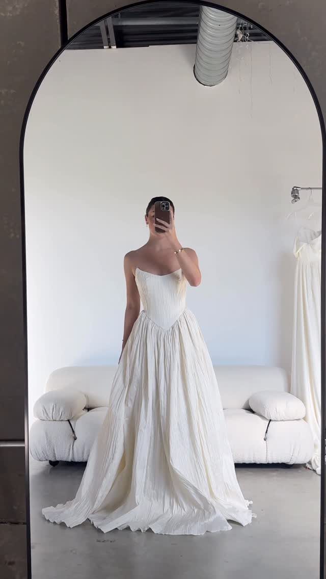 Person in a strapless off-white bridal gown taking a mirror selfie in a room.