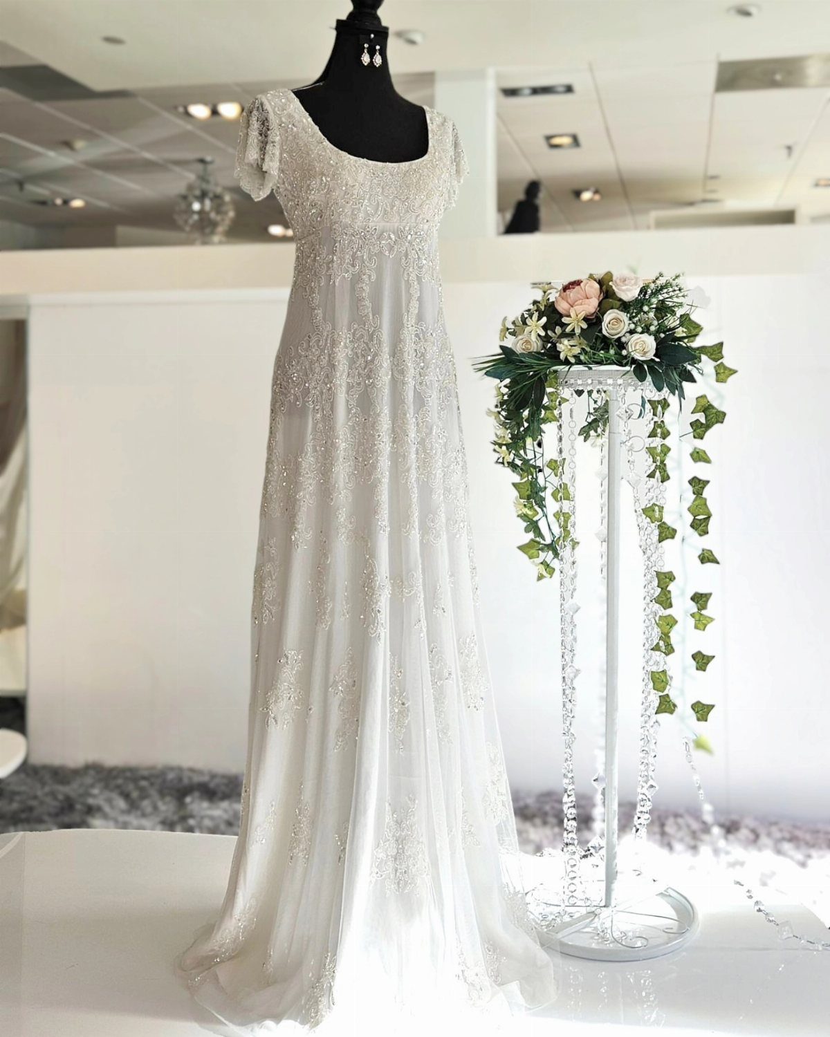 White embroidered wedding dress with empire waistline on a black mannequin next to flowers
