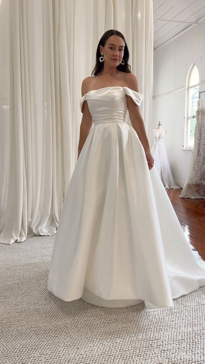 Woman wearing a white satin off-the-shoulder wedding gown in a bridal boutique.