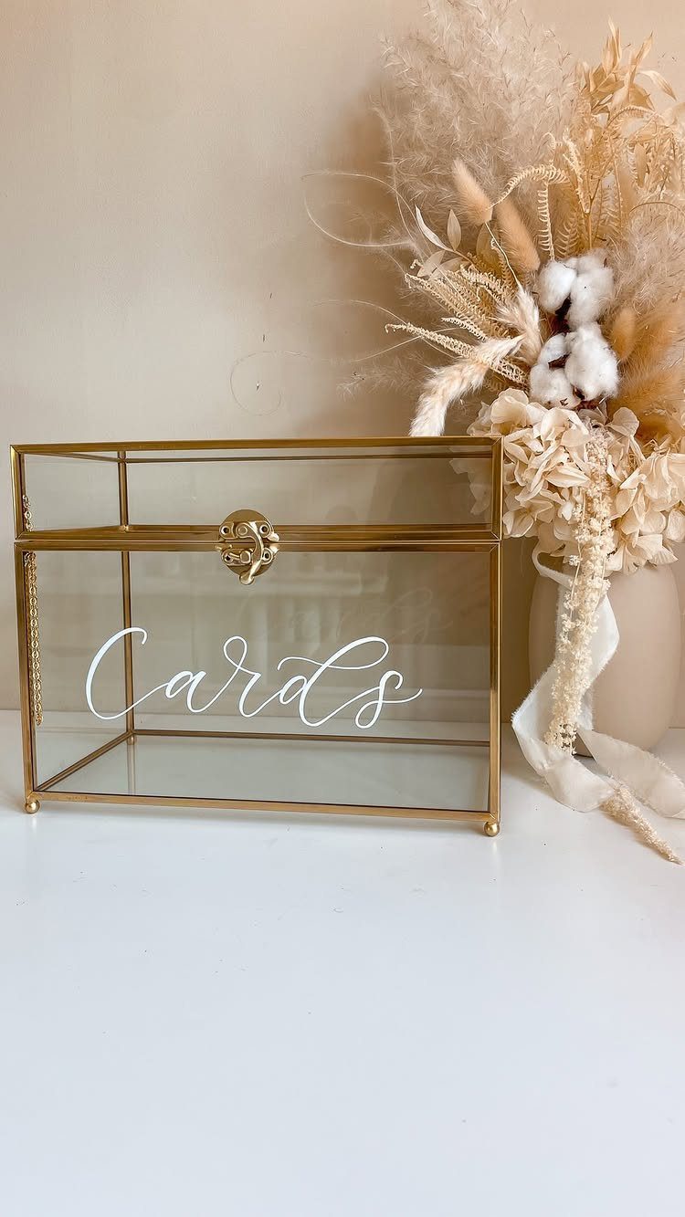 Gold glass card box with white cursive lettering next to dried floral arrangement