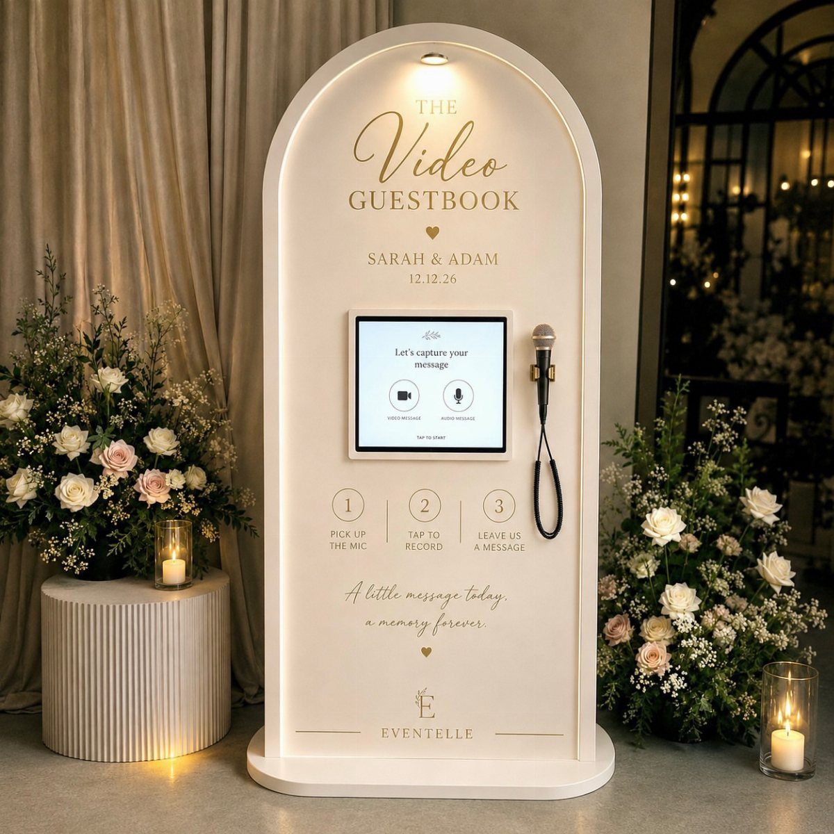 White arched video guestbook kiosk with a screen and microphone surrounded by floral arrangements.