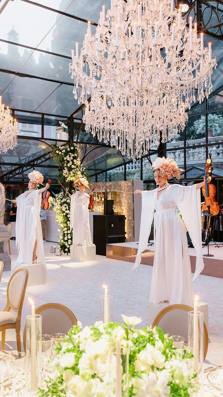 Musicians in white gowns performing with string instruments under a crystal chandelier
