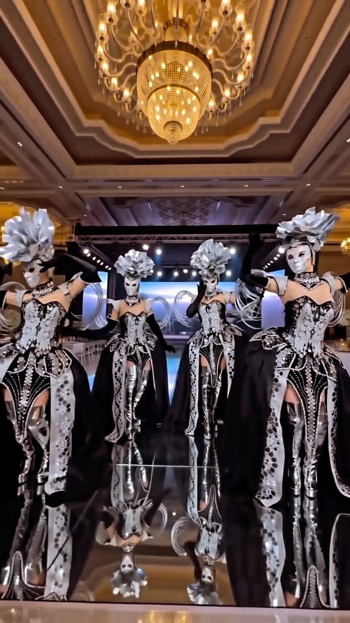 Performers in silver and black costumes posing in a luxury ballroom with chandeliers.