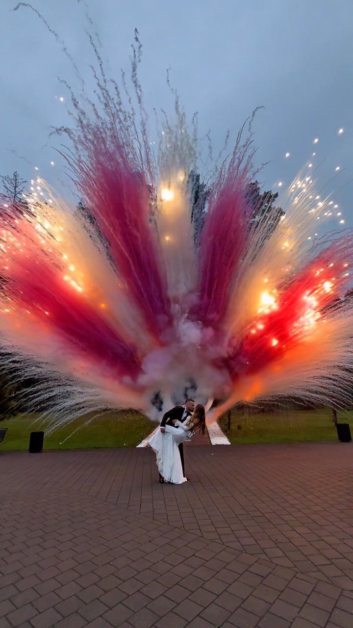 Bride and groom kissing in front of a colorful fireworks display at twilight.