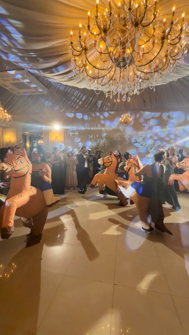 Guests in formal wear riding inflatable horses on a dance floor under a chandelier.