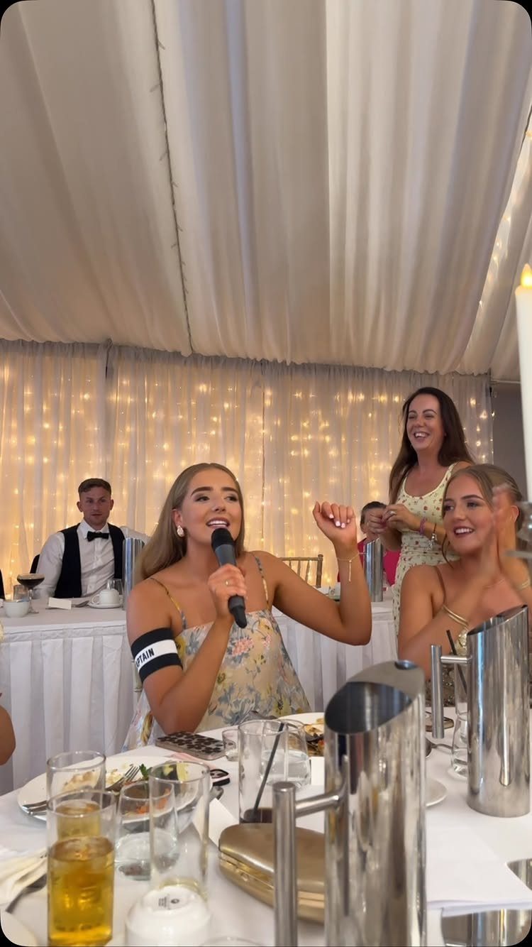Woman singing into a microphone at a wedding reception with guests and fairy lights.