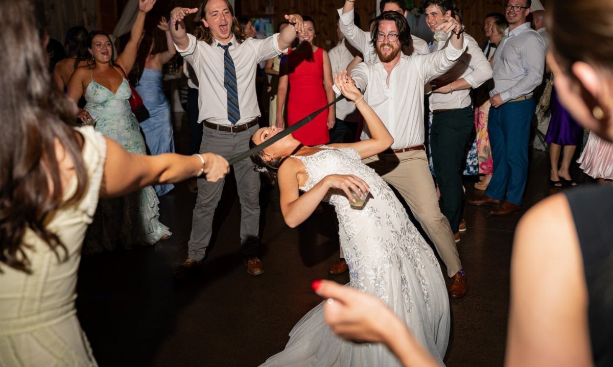 Bride in white gown leaning back to dance under a limbo stick at wedding