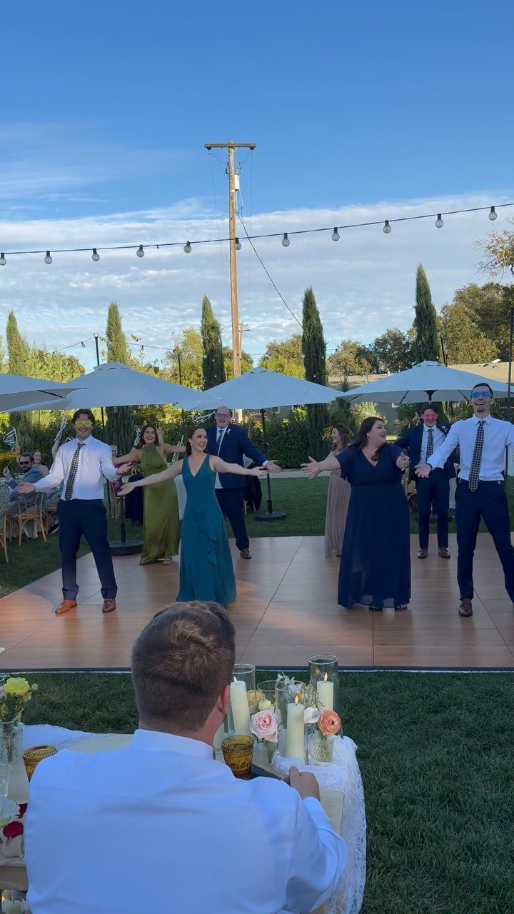 Group of formally dressed guests performing a choreographed dance on an outdoor dance floor.