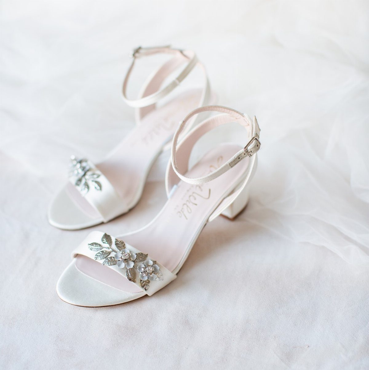 White satin block heel sandals with floral rhinestone details on white fabric.