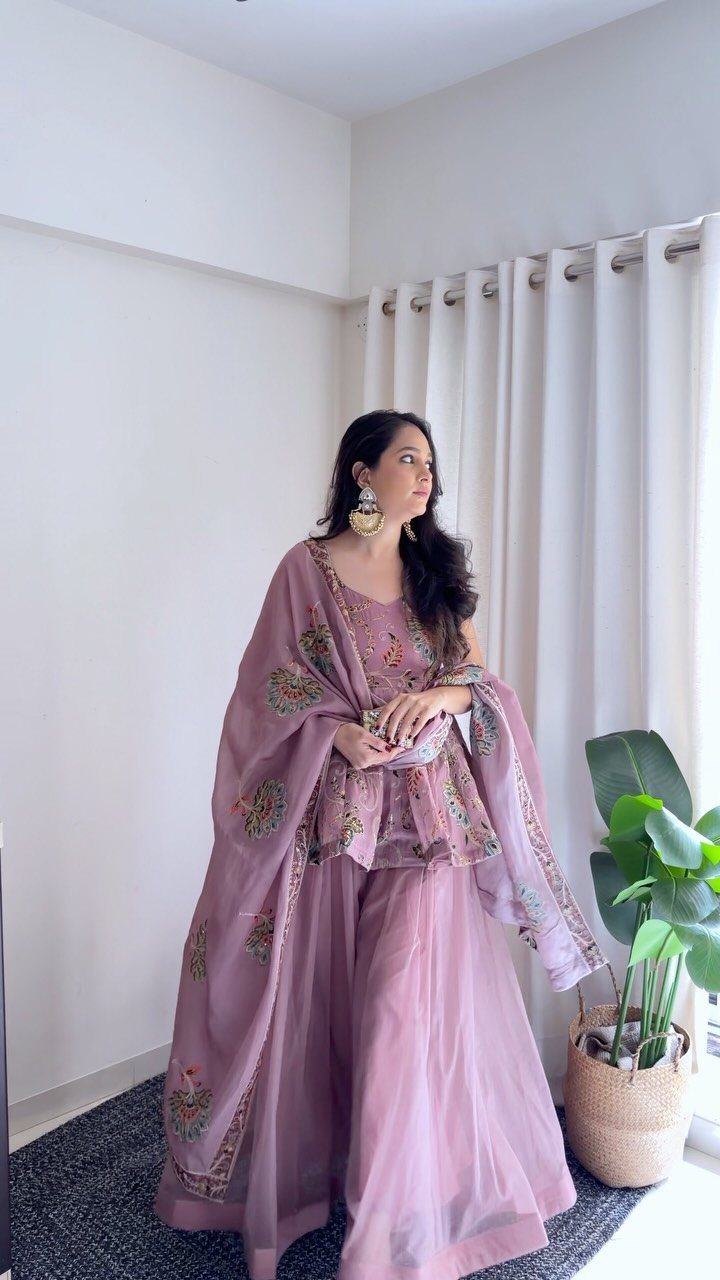 Woman in a mauve floral embroidered Indian outfit standing by white curtains.