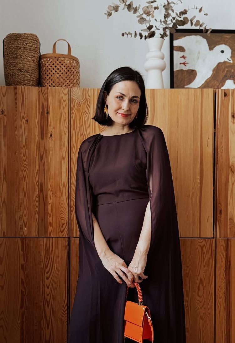 Woman in a flowing dark purple dress holding a small orange handbag indoors.
