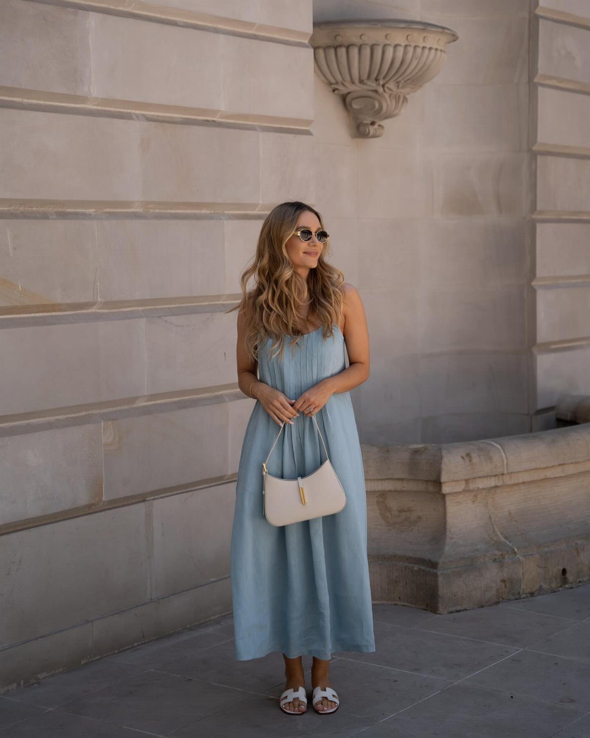 Woman wearing a light blue midi dress and white sandals against a stone wall.