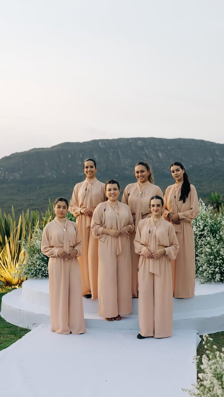 Six women in matching peach long-sleeved outfits posing on a white outdoor platform.