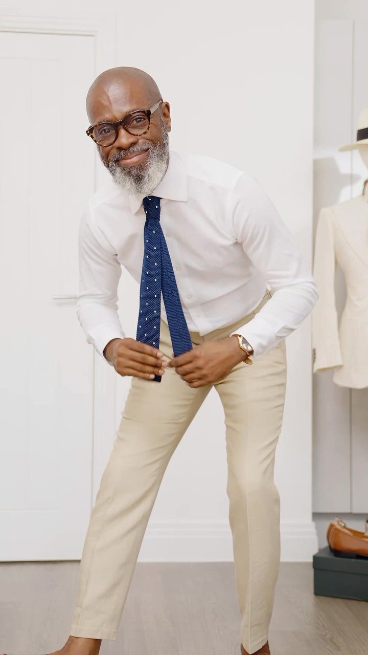 Man in white shirt, navy polka dot tie, and beige trousers posing indoors.