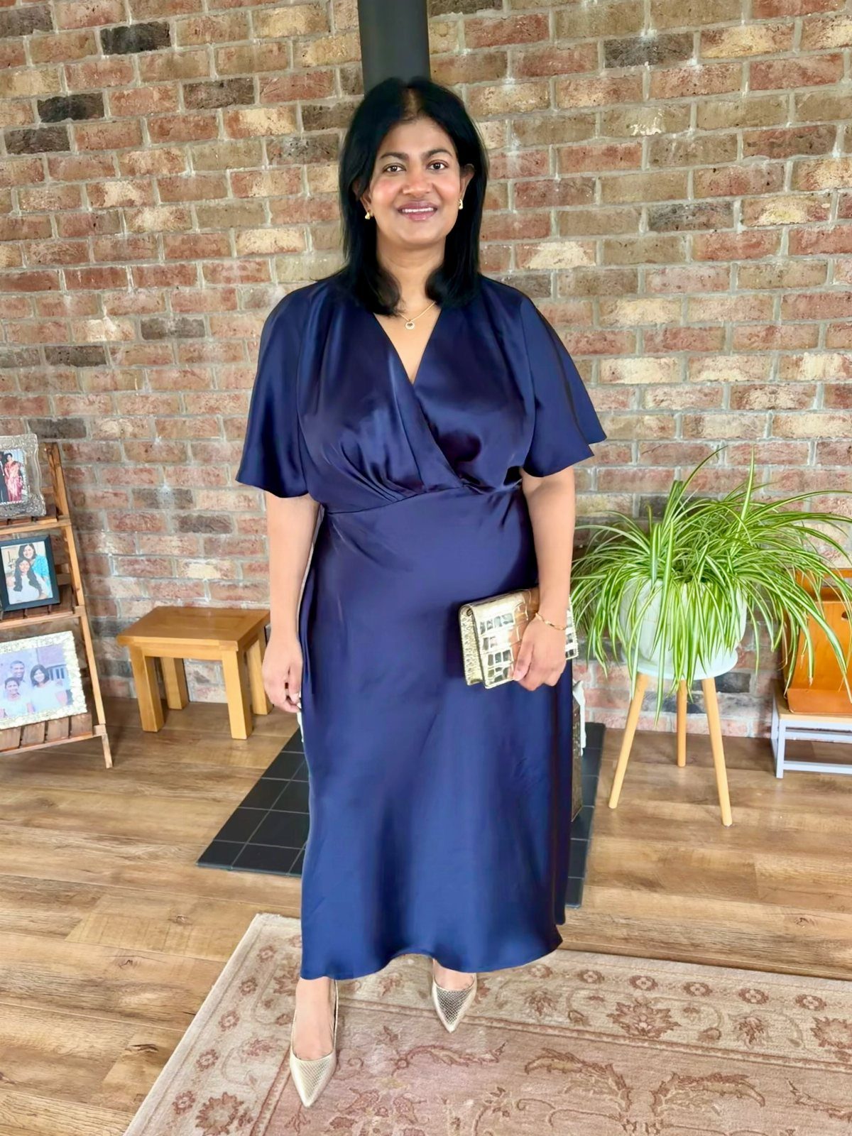 Woman in navy blue satin midi wrap dress with gold heels and clutch.