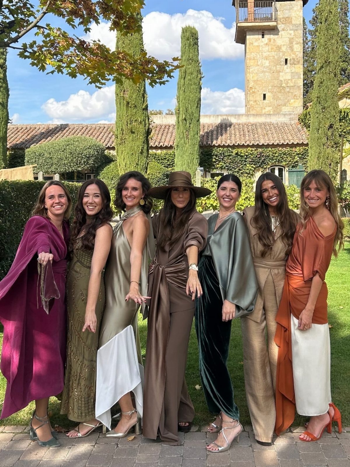Seven women in autumn-colored formal dresses and jumpsuits posing in a garden setting.