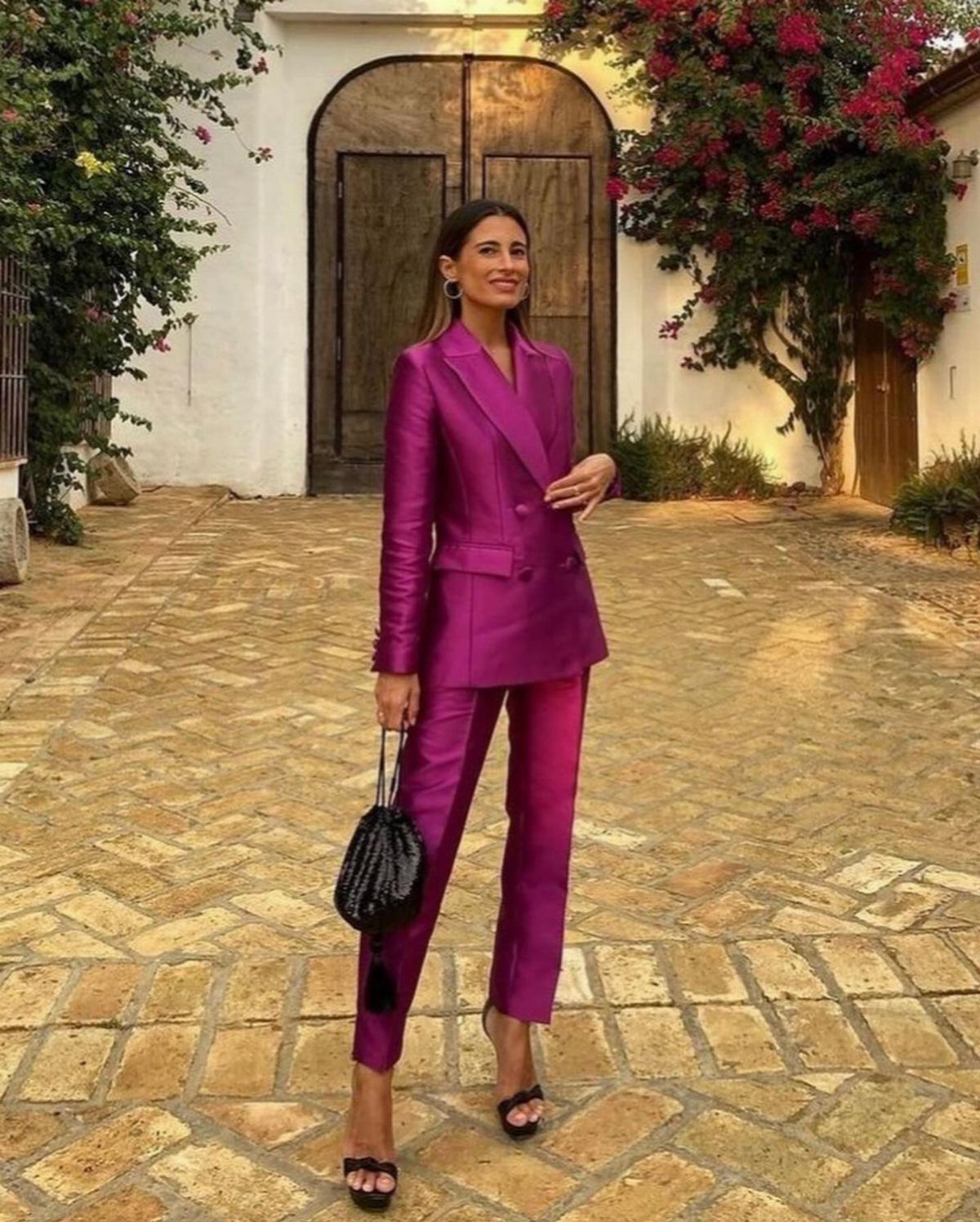 Woman in a magenta tailored pantsuit with black heels and handbag on cobblestones.