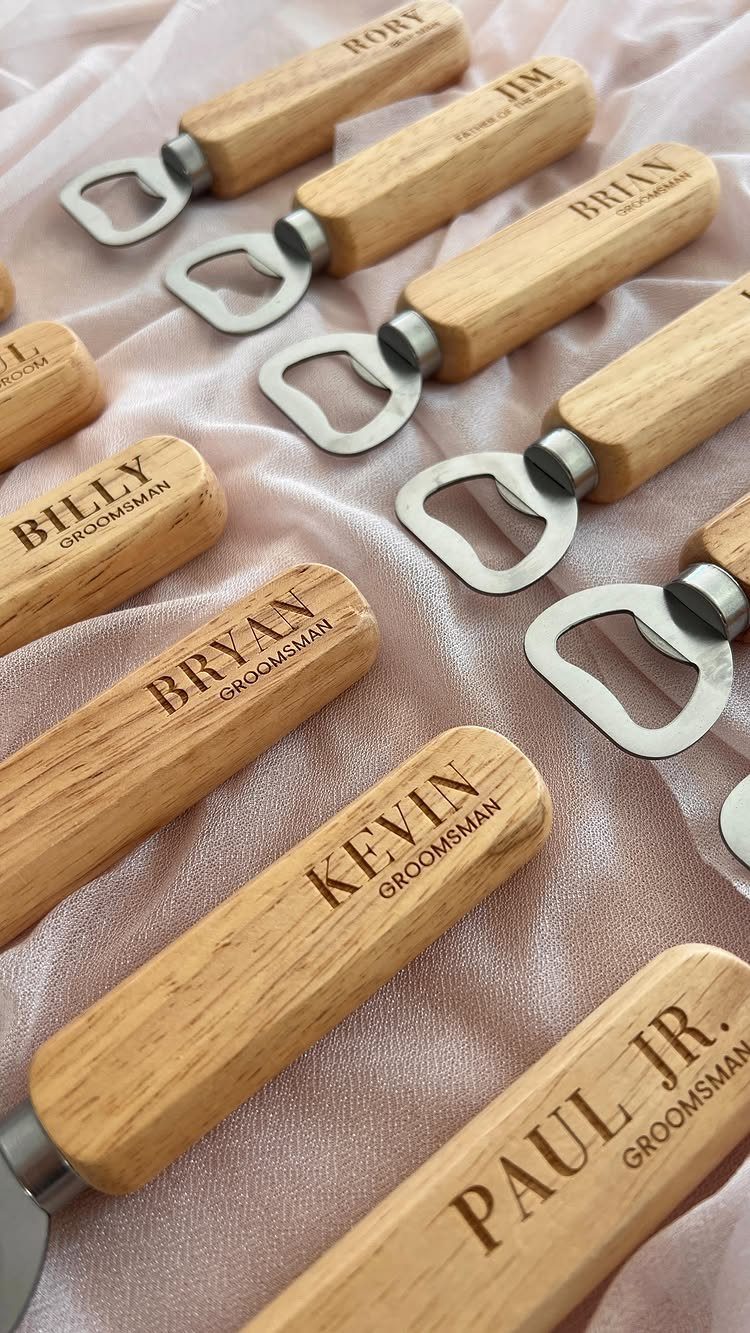 Engraved wooden bottle openers for groomsmen arranged on light pink draped fabric.
