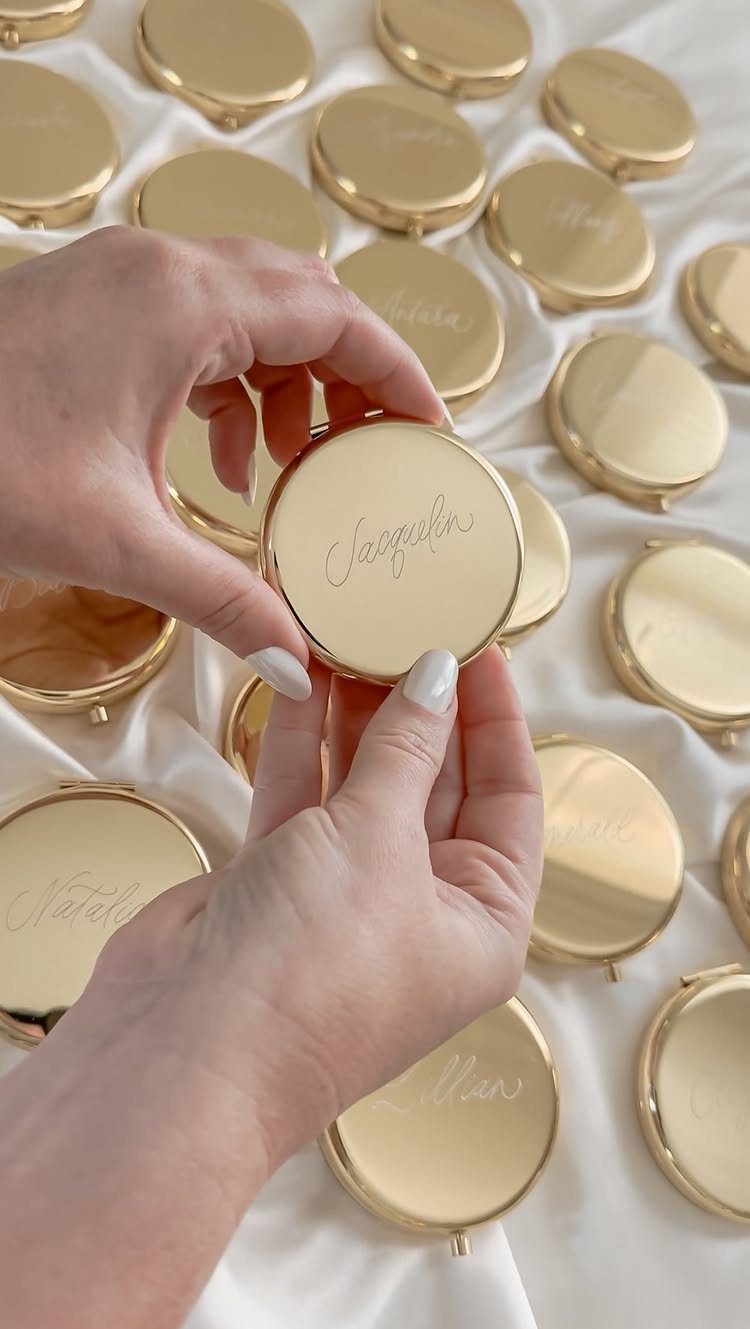 Hands holding a gold personalized compact mirror among many others on white fabric
