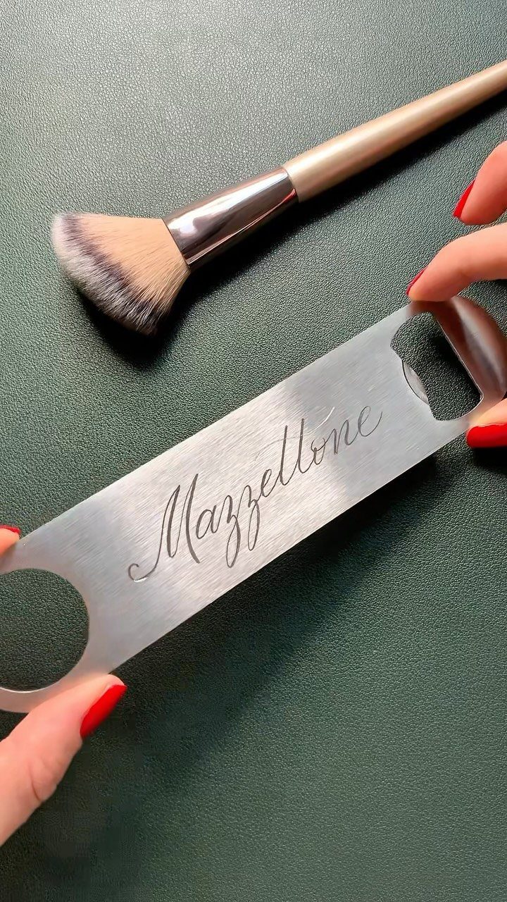 Engraved silver metal bottle opener held by hands with red nail polish.