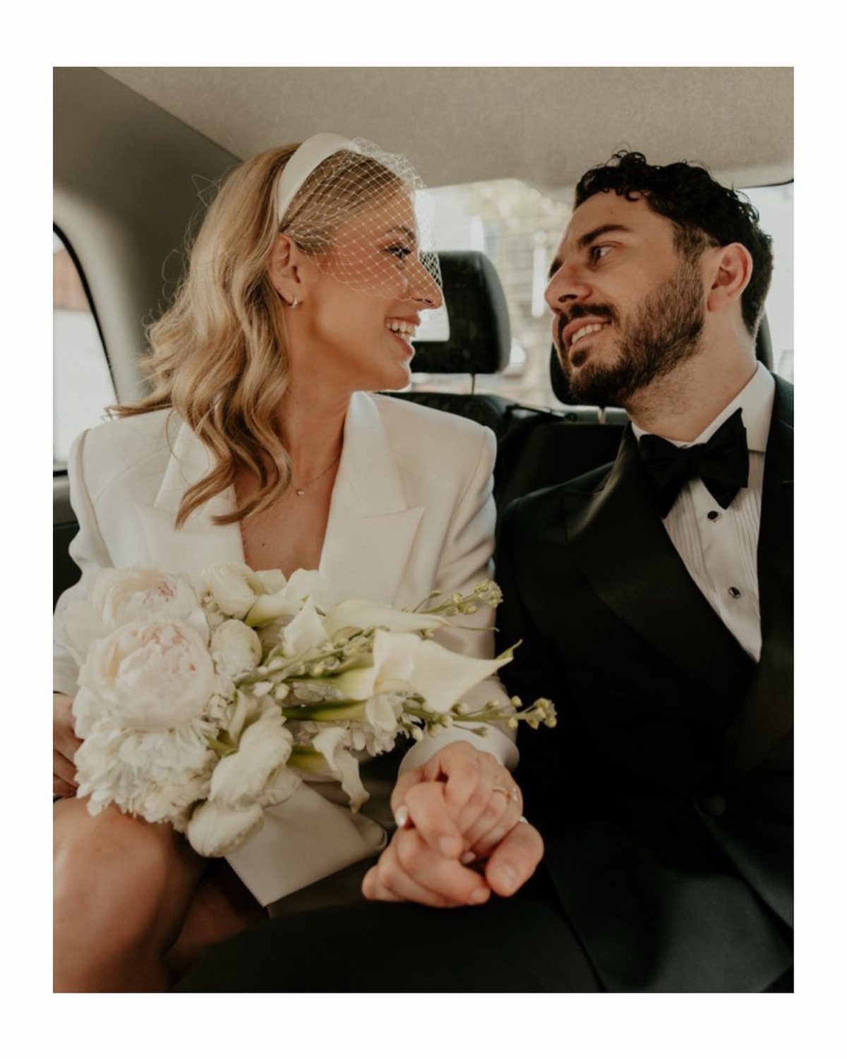 Bride with blonde waves and birdcage veil smiling at groom in a car