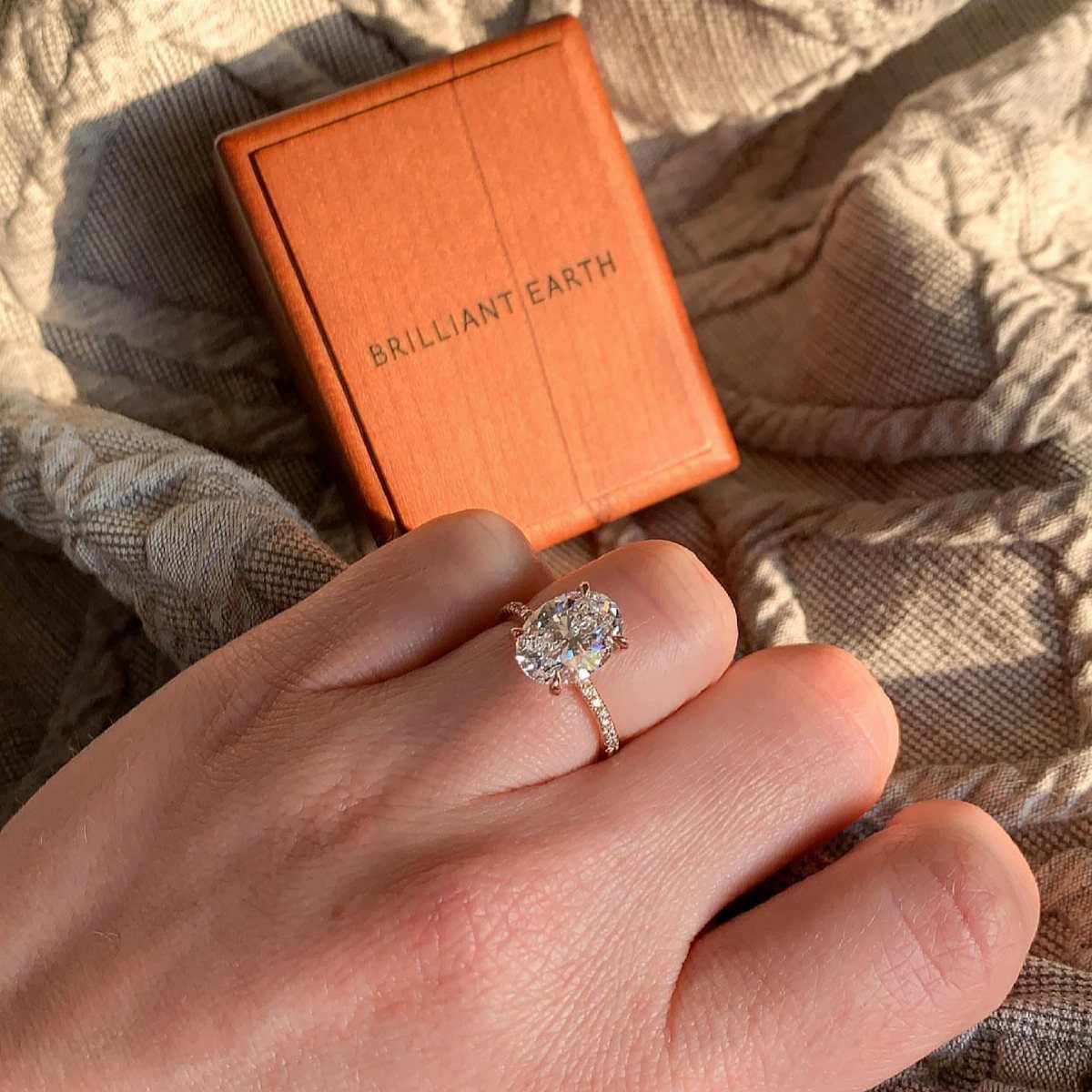 Diamond engagement ring on a finger with a Brilliant Earth jewelry box behind it.
