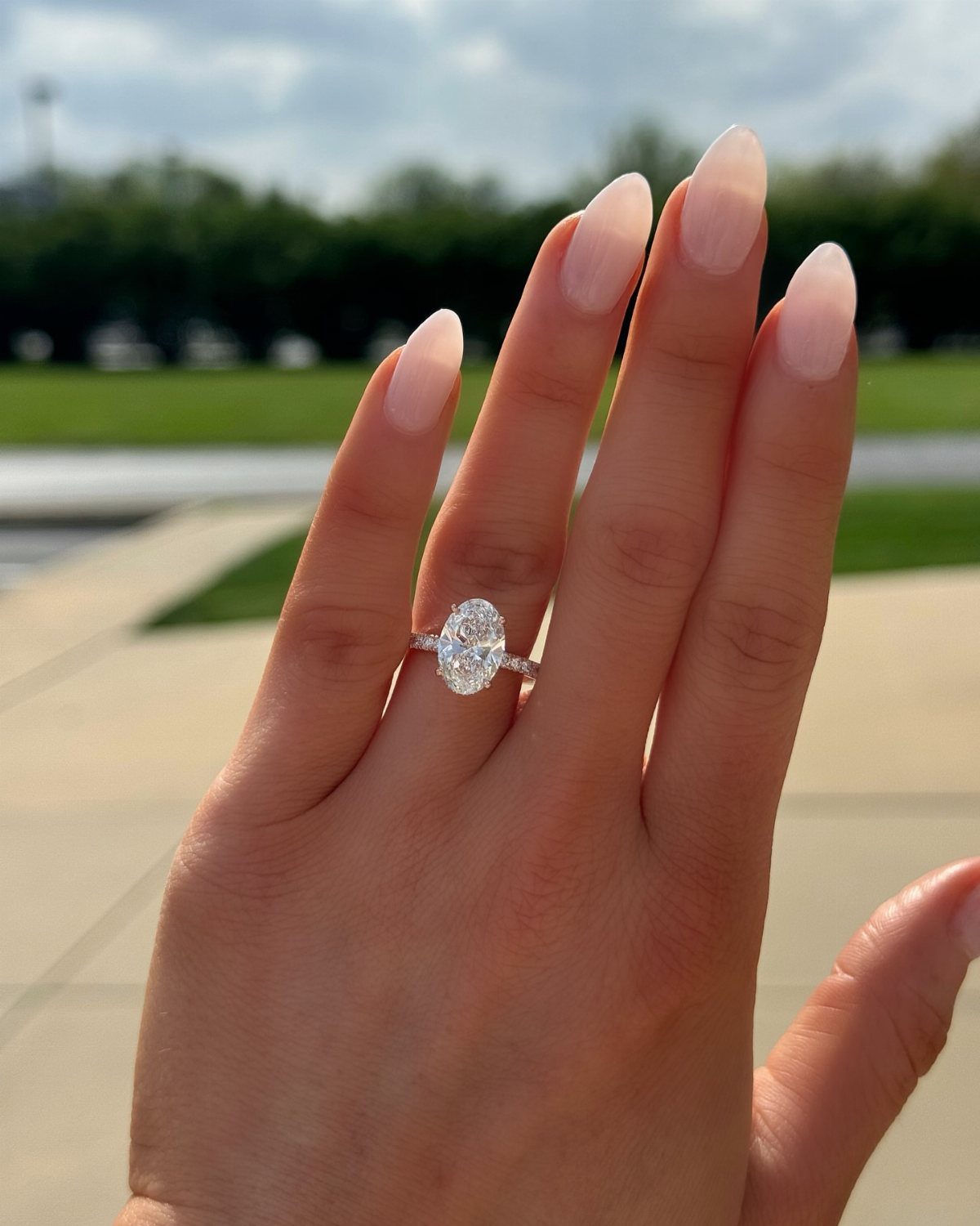 Oval-cut diamond engagement ring on a hand with neutral manicured nails outdoors.